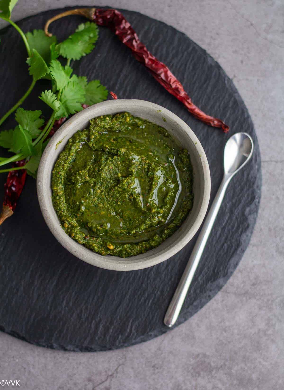 homemade cilantro shallots chutney in white bowl placed on black board with a spoon, dried chilies and cilantro on the side