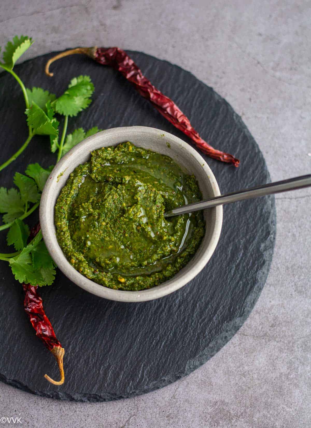 kothamalli chutney served in white ceramic bowl placed on black slate board with chilies and cilantro on the side