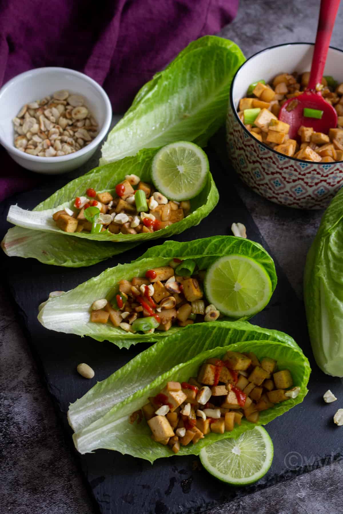 3 lettuce cups filled with tofu mixtures and served on a black slate board with some peanuts and the filling placed on a bowl on the side