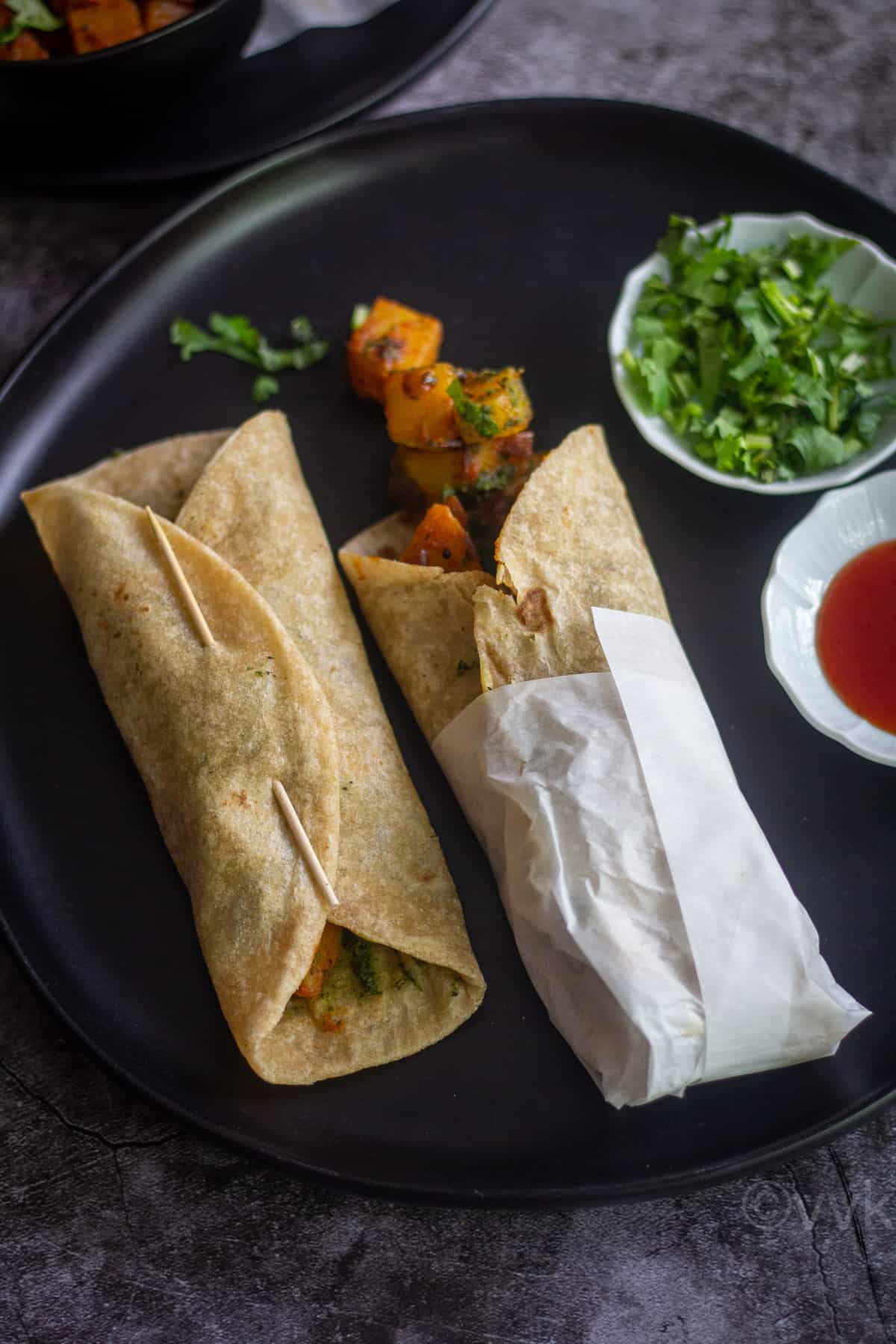 aloo kathi rolls served in black plate. One roll folded in parchment paper
