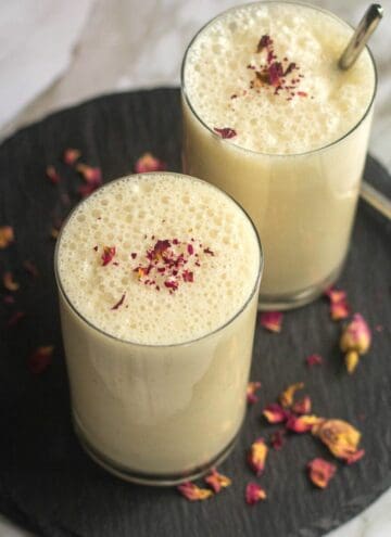 square image of gulkand lassi served in two glasses placed on black slate board