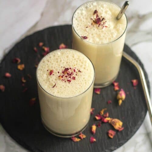 square image of gulkand lassi served in two glasses placed on black slate board