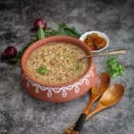 square image of gruel served in clay pot with pickle and other condiments on the side