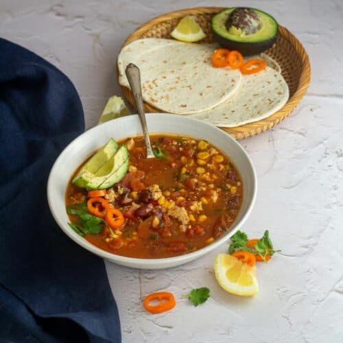 square image of taco soup served in white bowl with tortillas and avocado on the side