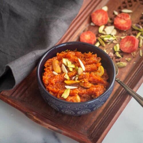 square image of carrot halwa served in blue bowl placed on wooden tray