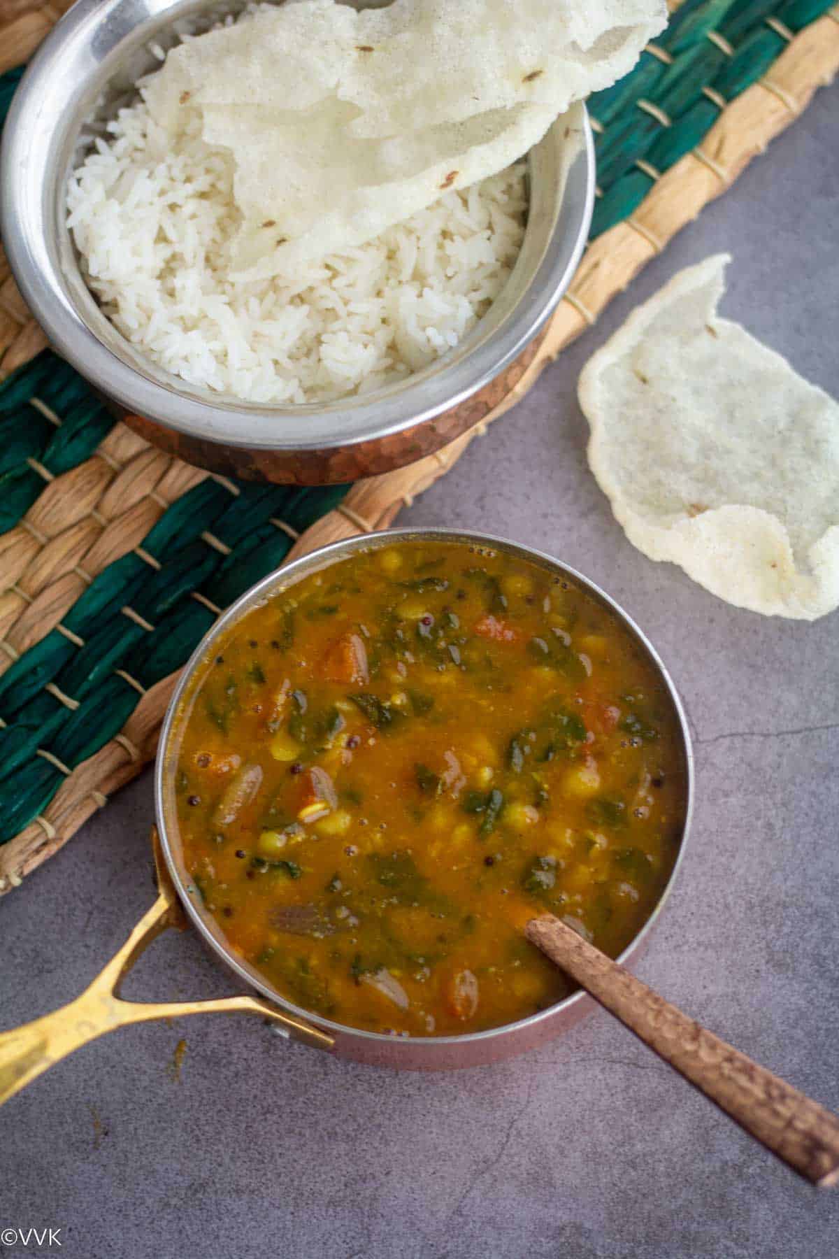 overhead shot of methi sambar served in brassware with rice and papad placed on green mat