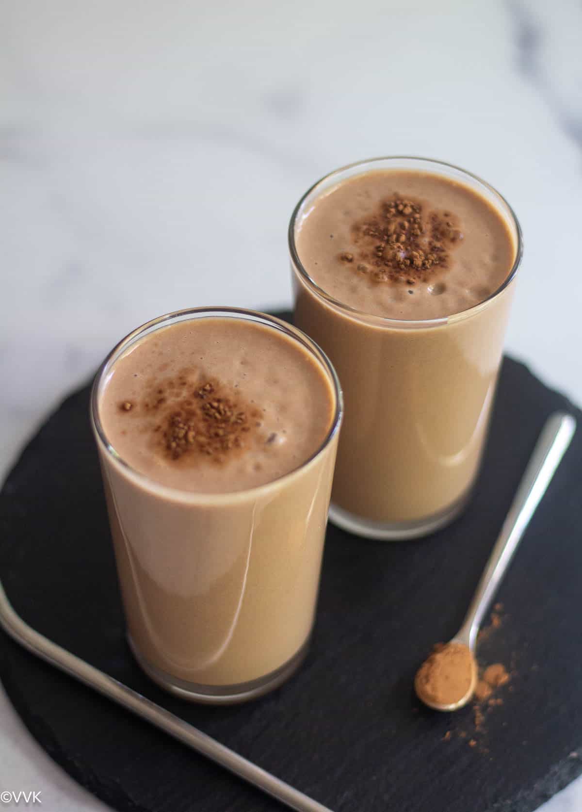 close up shot of chikoo lassi served in two glasses placed on black board