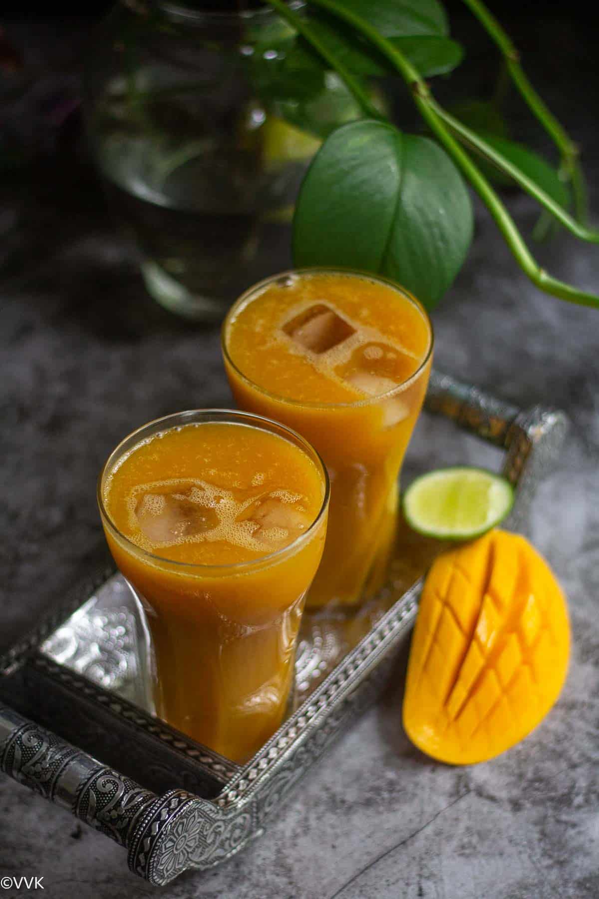 iced mango tea served in two tall glasses placed on a tray with mango and lemon wedge on the side