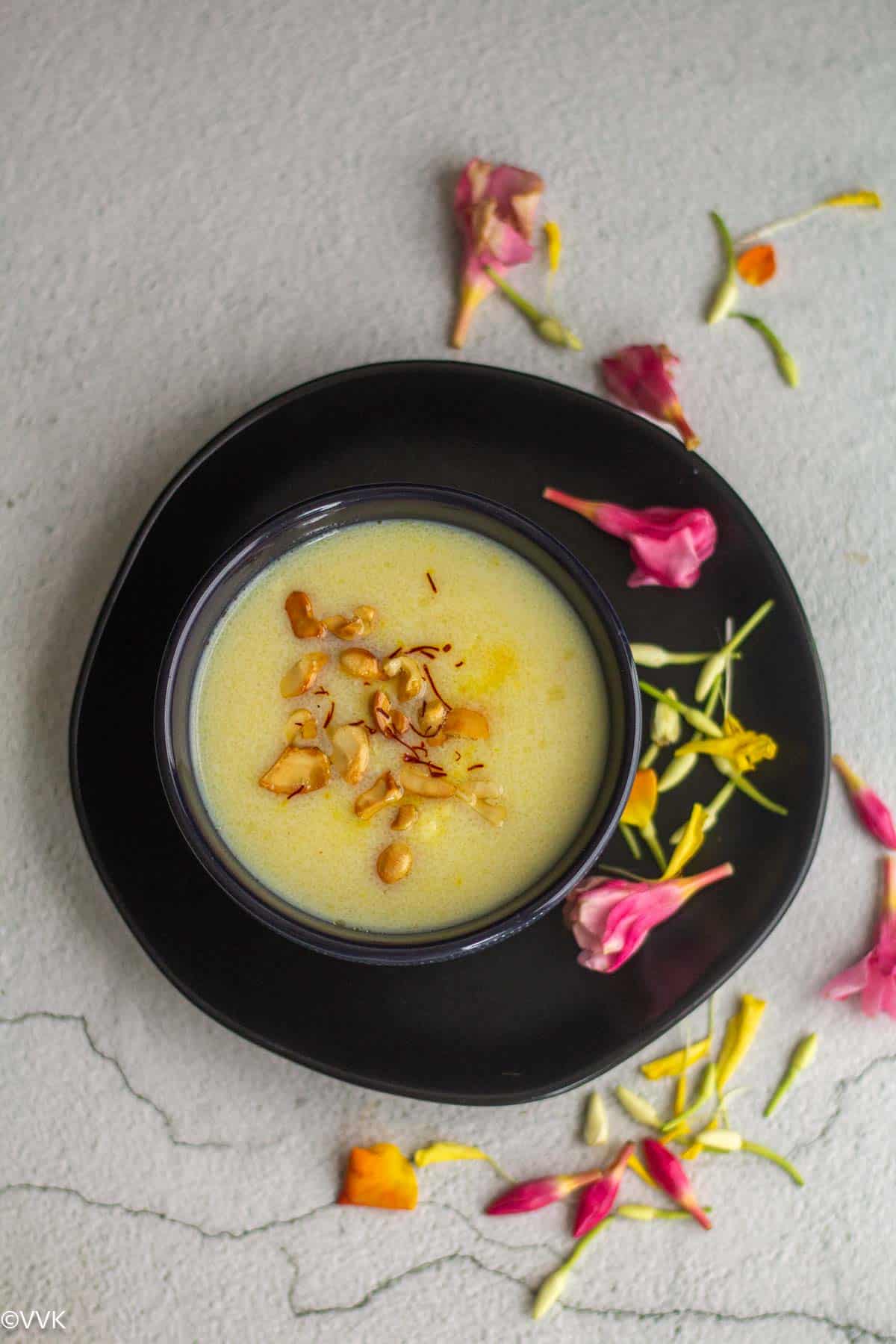 overhead shot of ravai payasam served on bowl placed on a black plate with flowers on the side