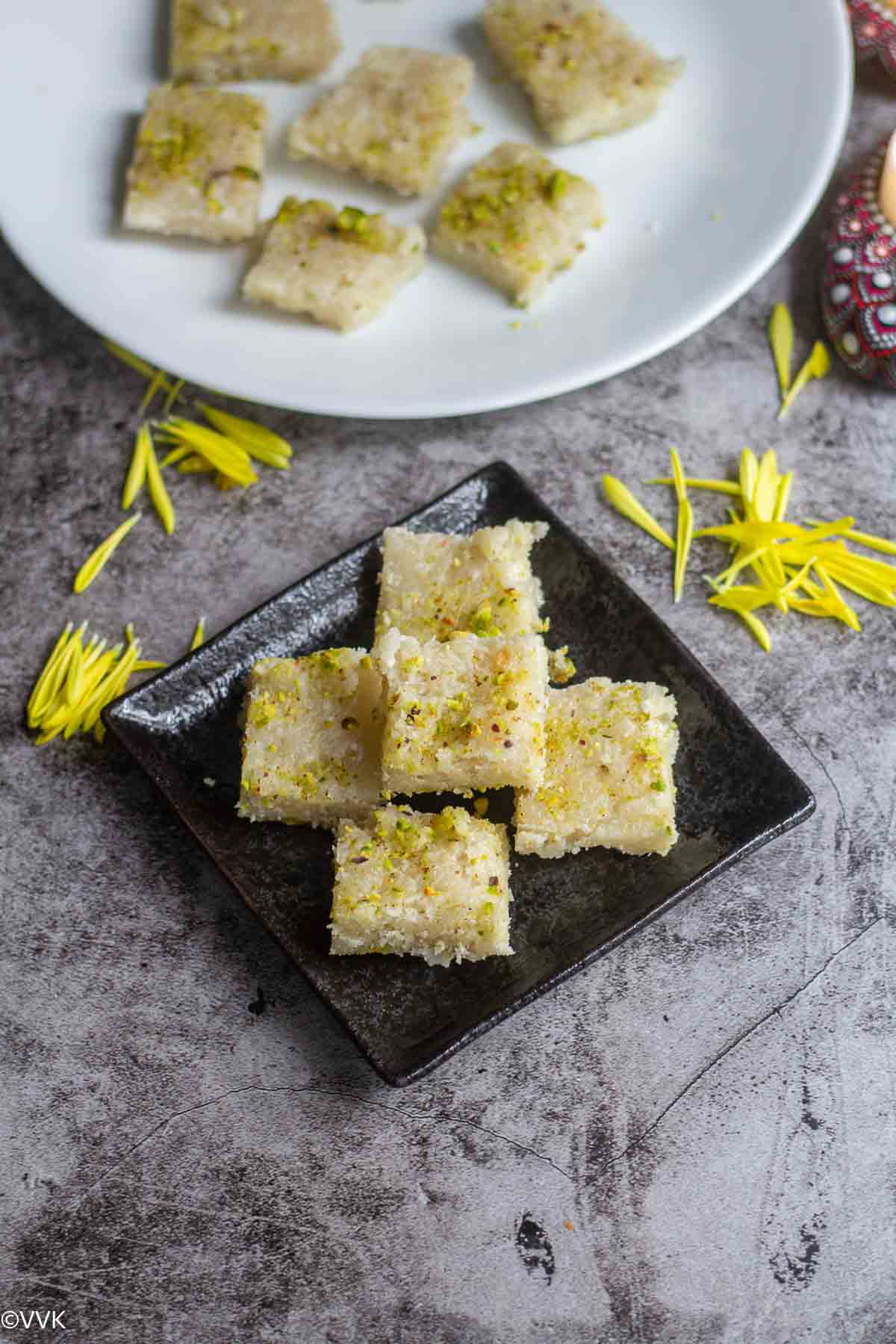 Thengai burfi in black plate and more in the back on white plate with yellow flowers drizzled