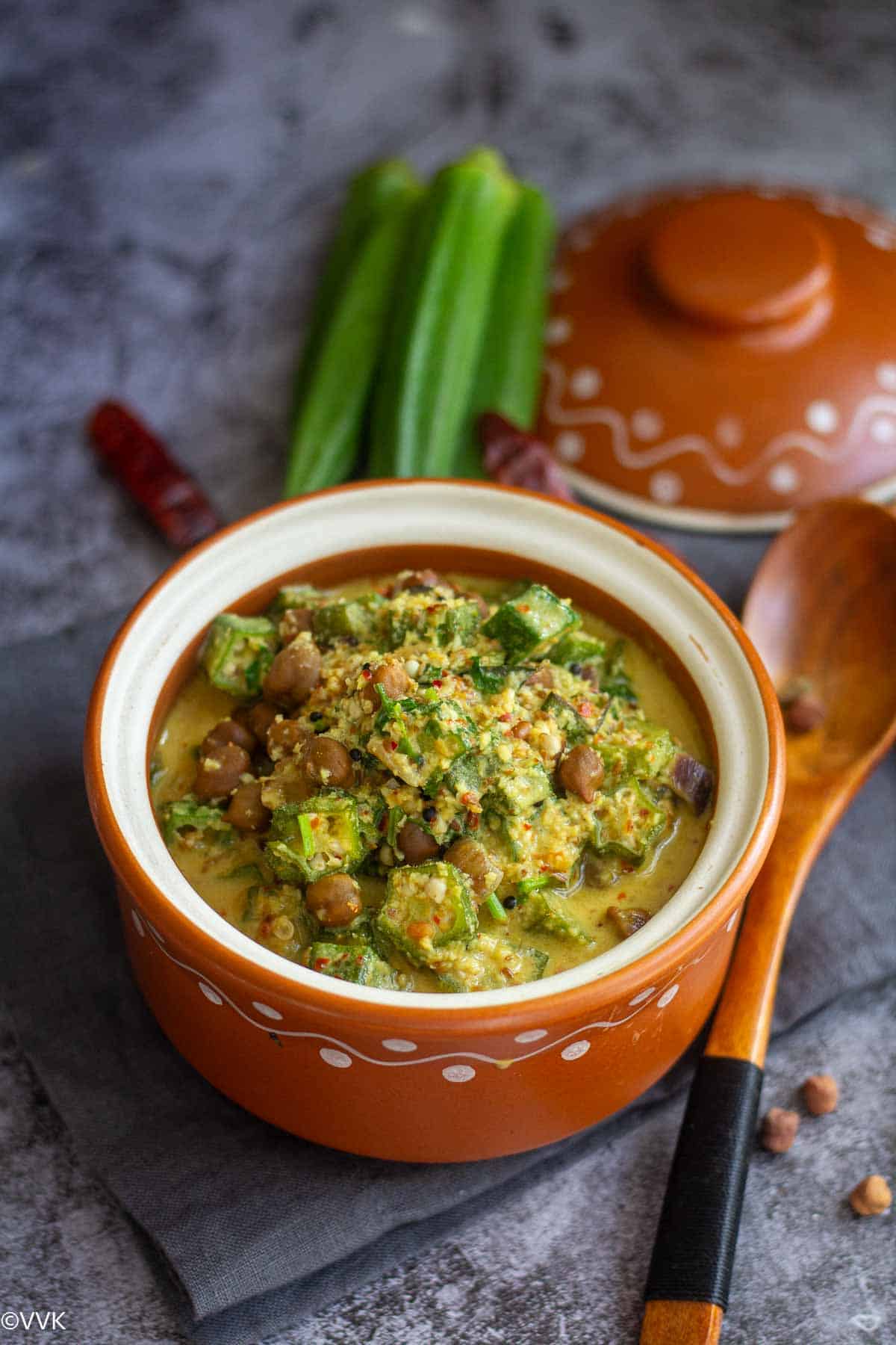 slanting shot of vendakkai kootu in clay bowl with some okra red chilies and ladle on the side