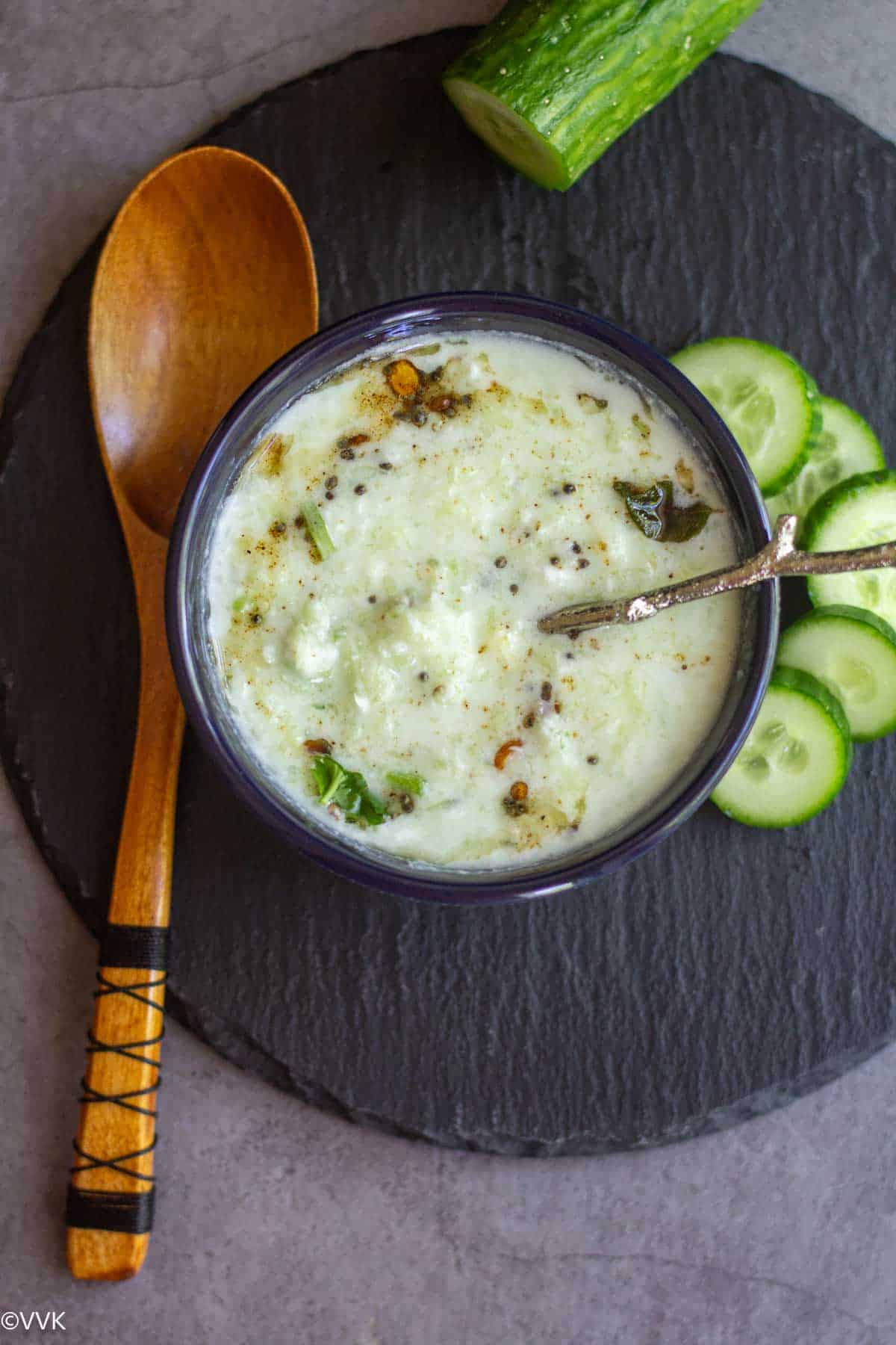 vellarikkai thayir pachadi served in a blue bowl with a spoon inside with cucumber slices on the side placed on black slate and a wooden spoon on the side